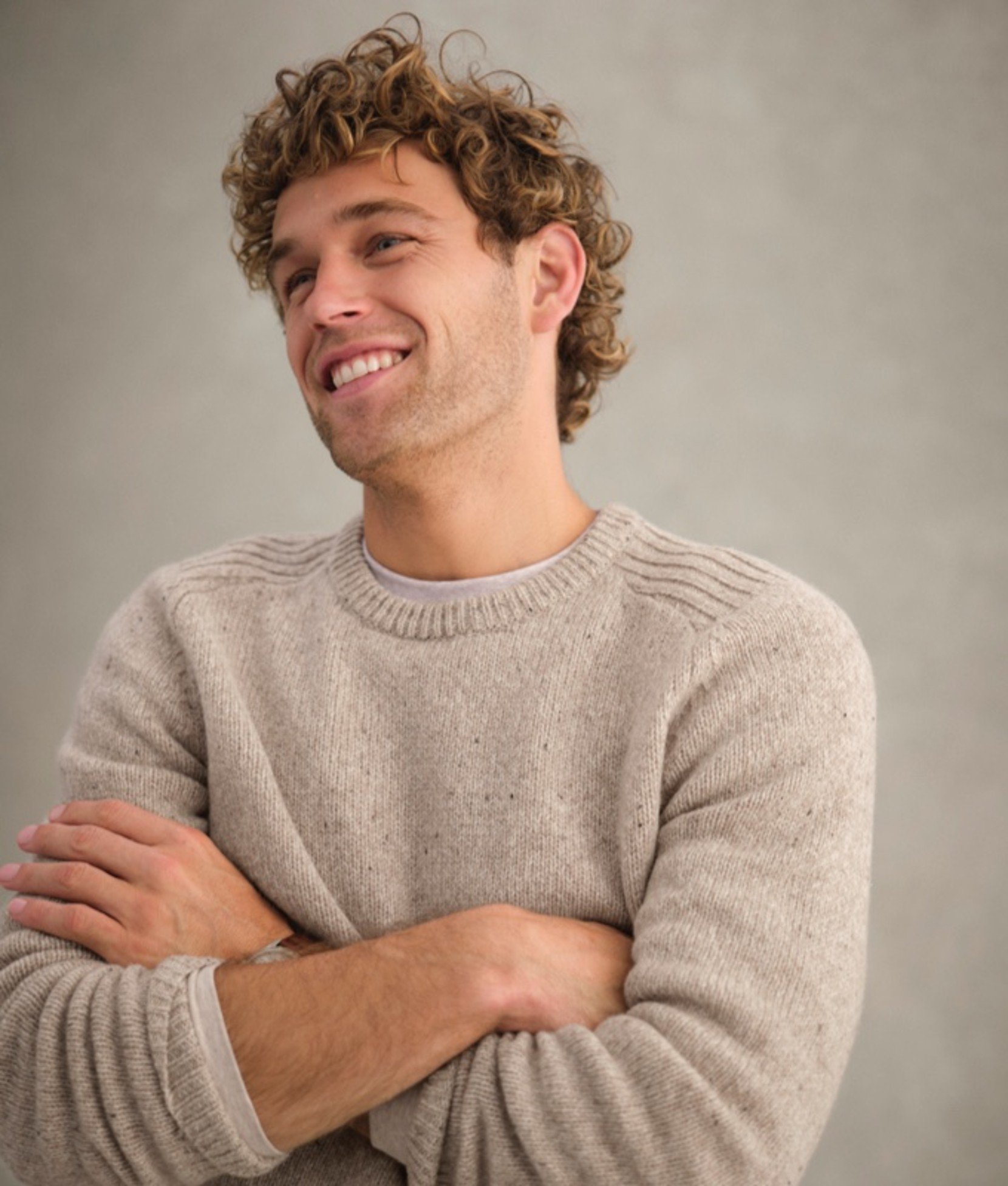 A man in beige knit sweater with arms crossed, neutral background.