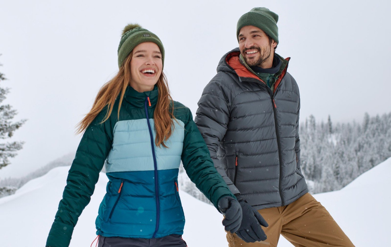 Two people in winter jackets and beanies walking through a snowy mountain landscape.