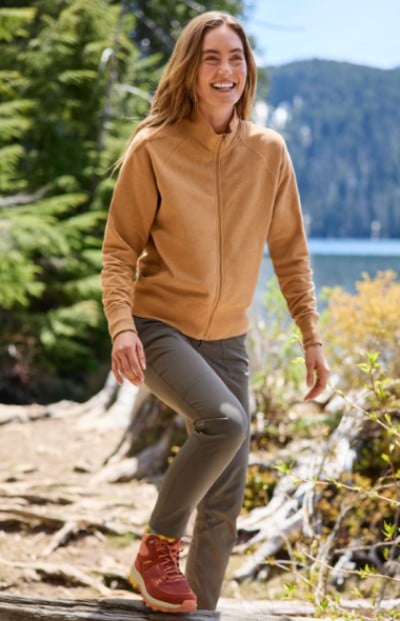 A woman in a beige jacket and grey pants walks on a forest trail by a lake. The scene is peaceful and natural. 