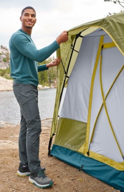 Person setting up a green and yellow tent by a lakeside, wearing a teal long-sleeve shirt, gray pants, and hiking shoes.