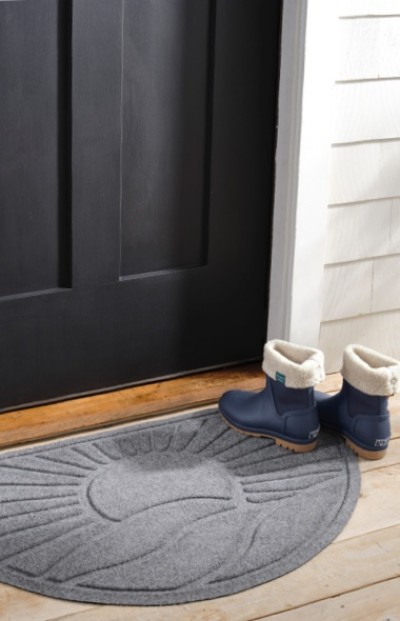Blue winter boots with white fur lining sit on a wooden porch floor beside a black door and gray semicircular doormat.