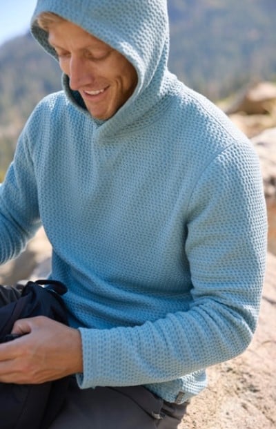 A man wearing a baby blue hooded sweatshirt stops on a trail to reach into his pack.