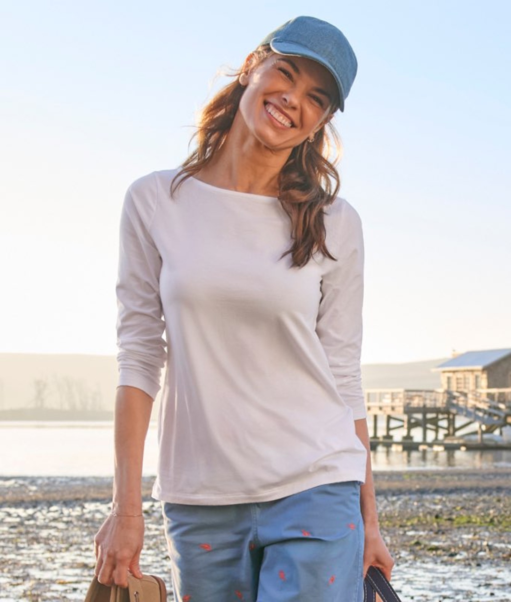 Person wearing a light long-sleeve top and cap, standing near the ocean on a bright day.