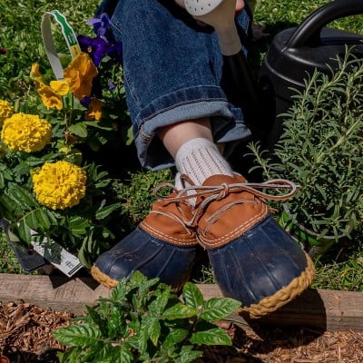 Person gardening in blue jeans and duck boots near yellow flowers.